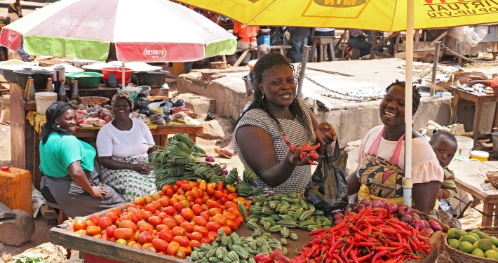 Street market scene in Nigeria with fresh produce laid out on tables, and a number of woman sitting around