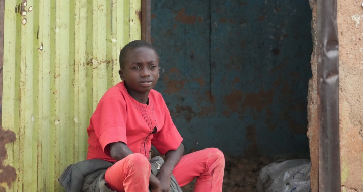 A young boy dressed in red, sitting in front of a corrugated iron structure